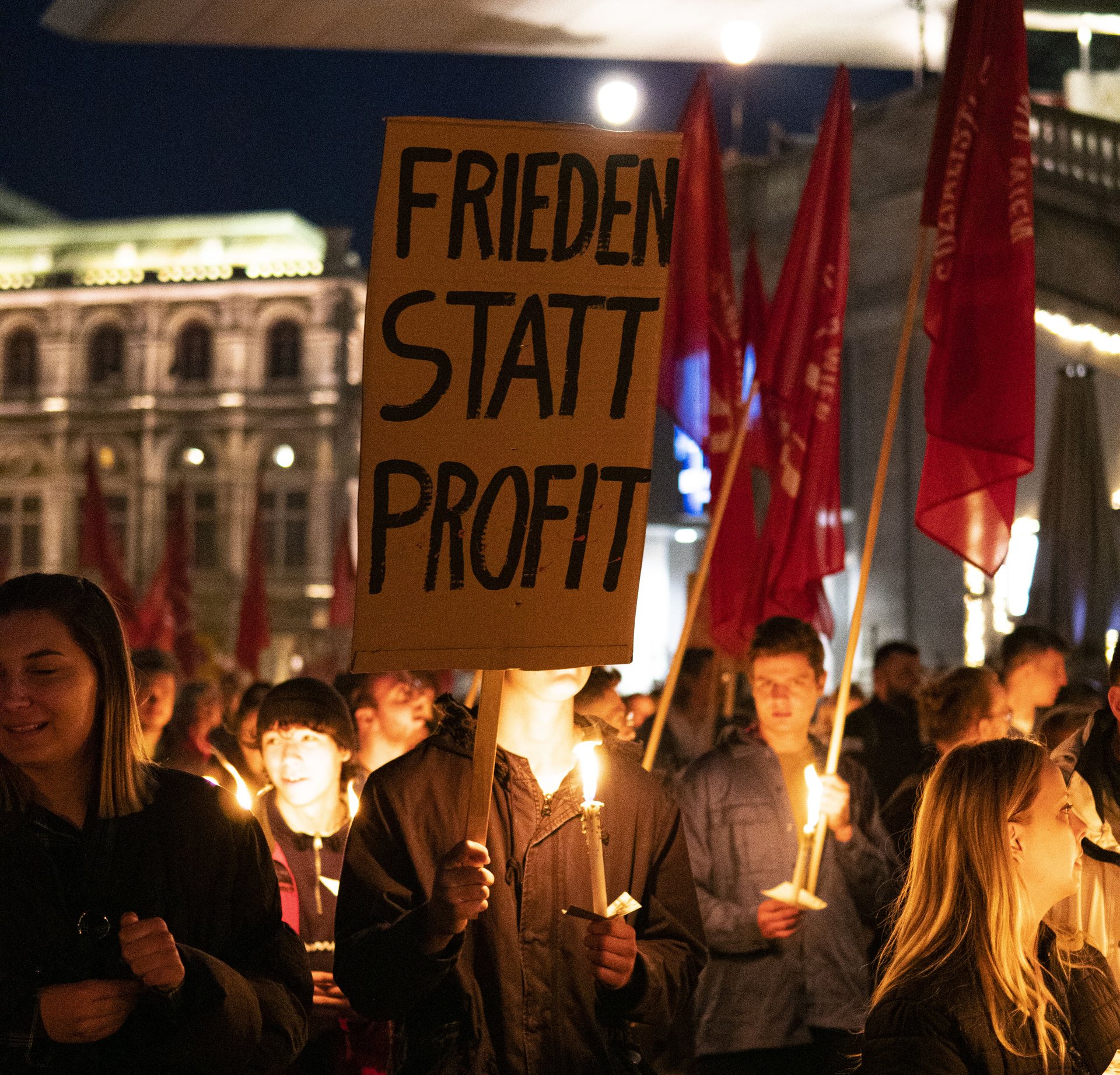 Fackelzug der SJ Wien. Fokus des Bildes liegt auf einem Schild, auf dem steht oben: Frieden statt Profite. Personen im Hintergrund halten Fackeln.