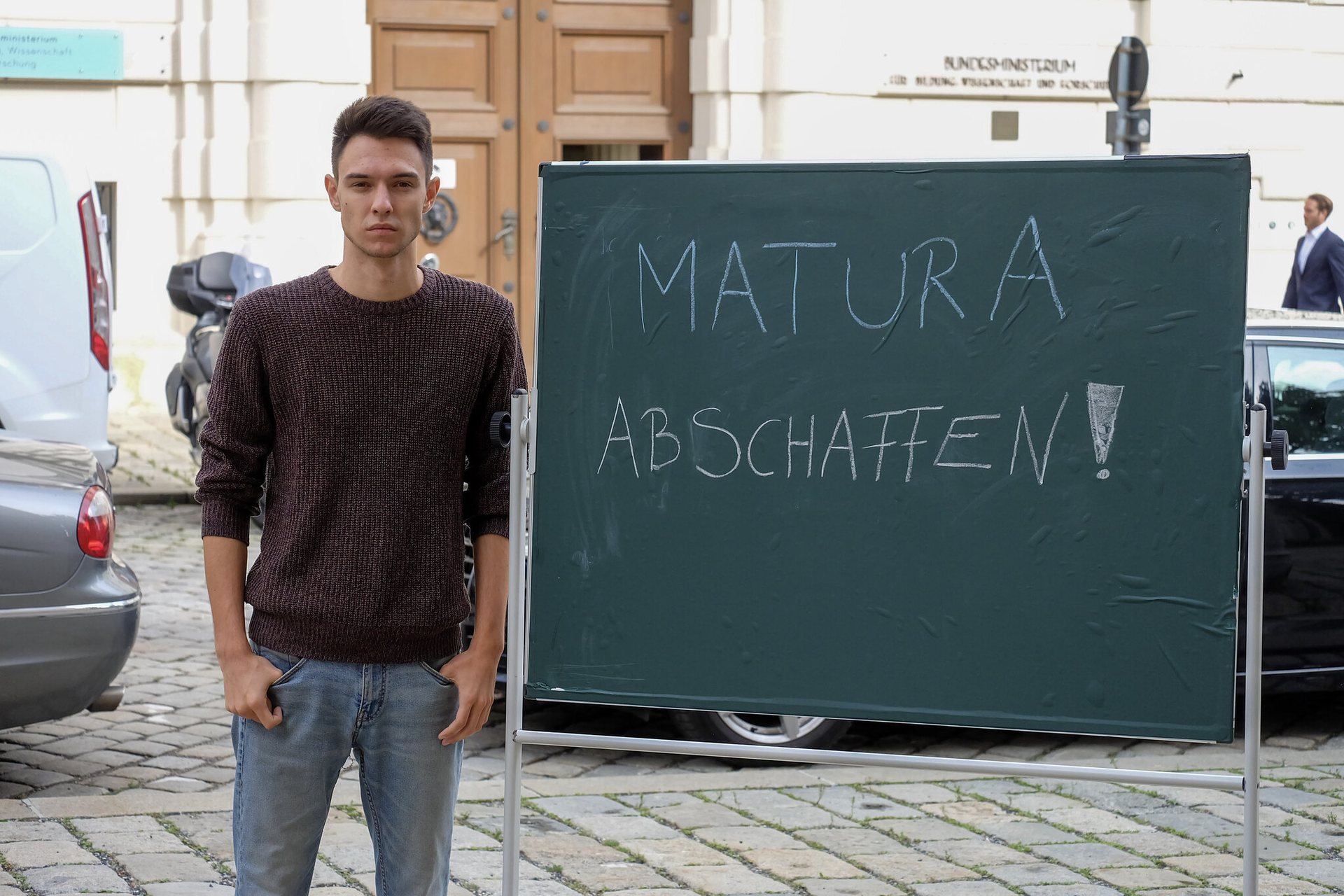 Paul Stich steht neben einer Tafel auf der oben steht "Matura abschaffen!"