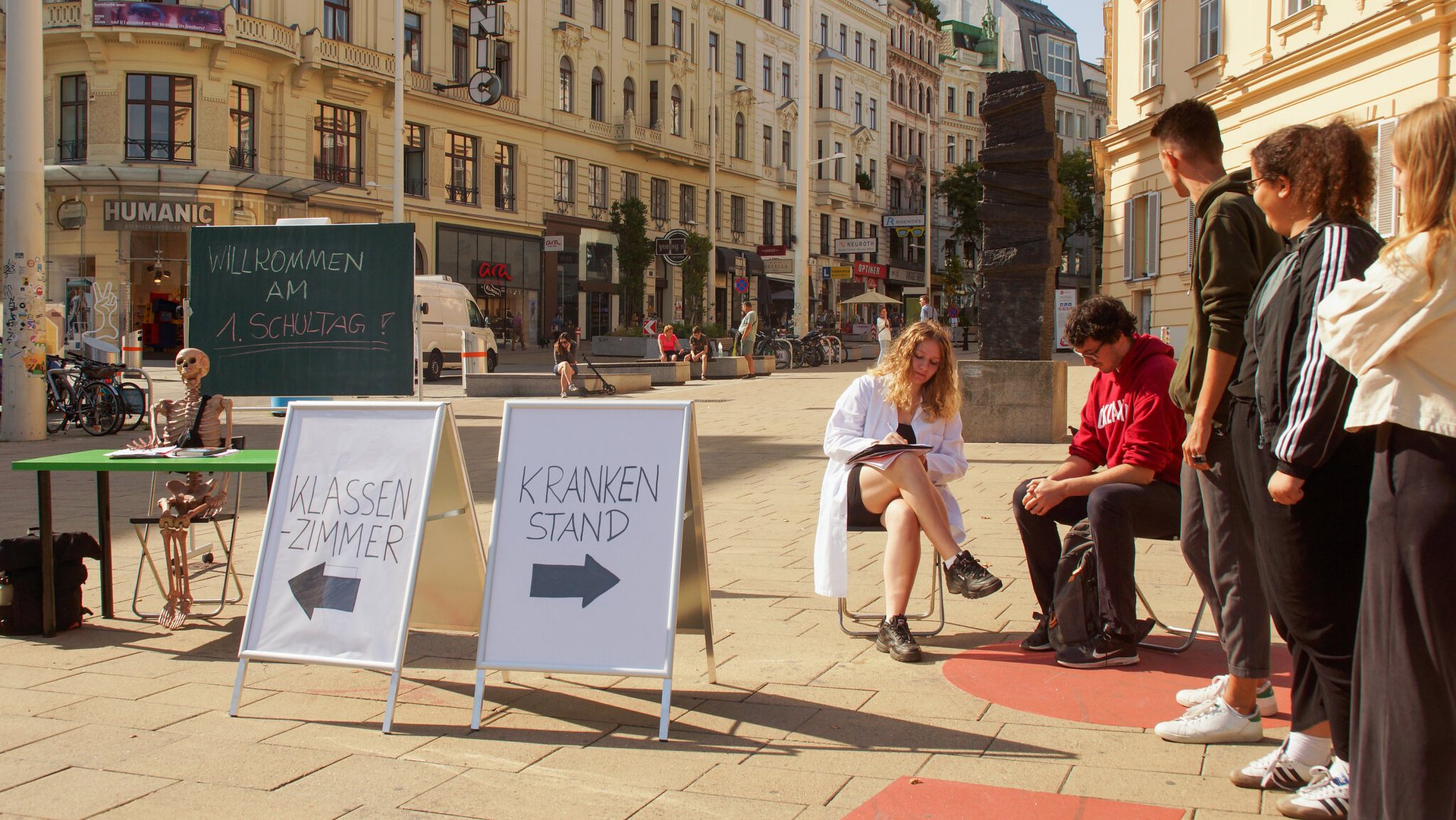 Eine Schulklasse ist am Platz der Menschenrechte in Wien aufgebaut. Aus zwei A-Ständern sind zwei Wege angeschrieben. Der erste Weg führt in ein Klassenzimmer, wo ein Skelett vor einer Tafel sitzt auf der oben steht "Willkommen am 1.Schultag!". Auf dem zweiten A-Ständer steht "Krankstand" angeschrieben, wo mehrere Jugendliche in einer Reihe stellen und darauf warten von der Ärztin krankgeschrieben zu werden. 