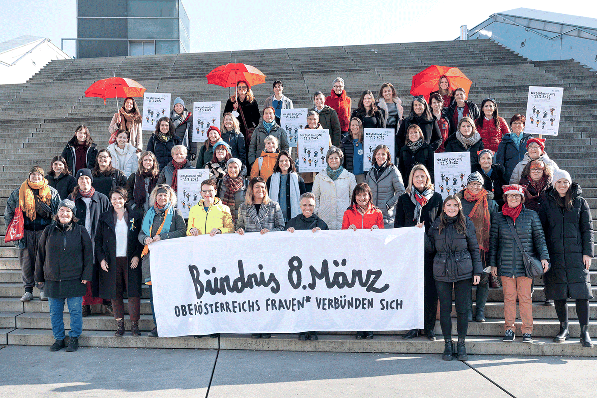 Dutzende Frauen stehen auf breiten Stufen in mehreren Reihen nebeneinander. In der ersten Reihe halten sie ein Banner auf der "Bündnis 8. März" steht.