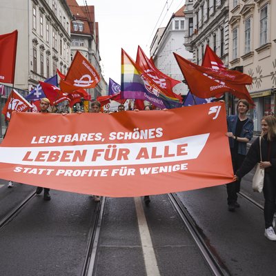 Demonstration. Mehrere Fahnen sind zu sehen, darunter eine Regenbogenfarbe. Auf dem vordersten Transparent des Demozuges steht: "Leistbares, schönes Leben für alle, statt Profite für wenige".