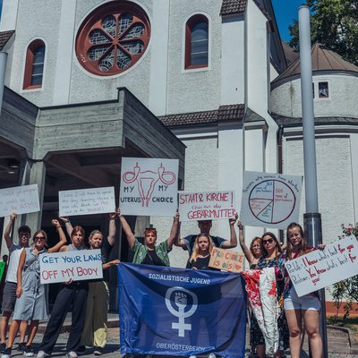 Frauen stehen vor einer Kirche und schauen wütend. Sie halten Schilder in die Höhe die das Selbstbestimmungsrecht von Frauen unterstreichen zB.: "Get your laws off my body", "My uterus, my choice"