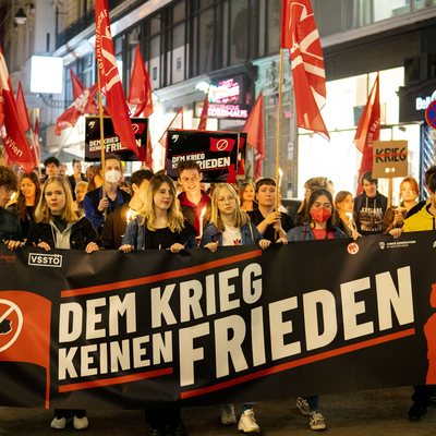 Fackelzug der Sozialistischen Jugend Wien. Der Demozug hält ganz vorne ein Transpi, auf dem steht oben "Dem Krieg keinen Frieden". Personen sind von vorne zu sehen.