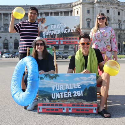 Vier Jugendliche sind sommerlich gekleidet und halten zwei Schilder: "Gratis Sommerticket - Für alle unter 26!"