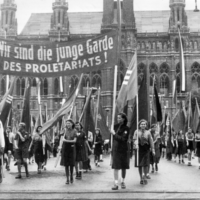 Jugendliche gehen in zwei Reihen hinterinander und halten Fahnen. Auf einem Flugtransparent ganz vorne steht oben: Wir sind die junge Garde des Proletariats! Zu sehen von vorne