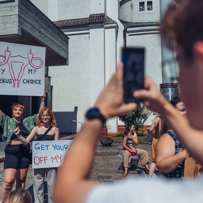 Eine Person ist verschwommen im Vordergrund, welche gerade ein Foto von zwei Frauen mit Schildern macht. Auf den Schildern ist "My Uterus, My Choice" zu lesen. Das Foto wurde auf der Kundgebung gegen Fundamentalist*innen gemacht.
