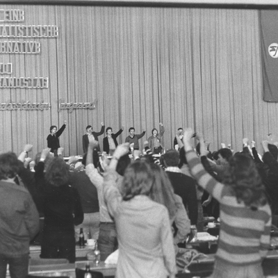 Ein schwarz-weiß Foto des 18. ordentlichen Verbandstages der Sozialistischen Jugend Österreich. Die Delegierten stehen mit erhobener Faust. Die Gruppe ist von hinten zu sehen, auf der Bühne stehen drei Leute.