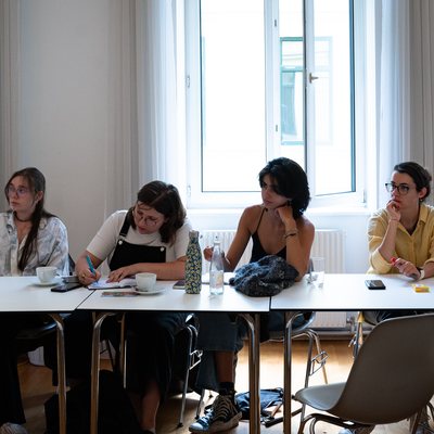 Fünf Frauen sitzen am Tisch und haben ihren Blick auf eine Person gerichtet die nicht zu sehen ist. Eine Frau schreibt etwas in einem Notizbuch mit.