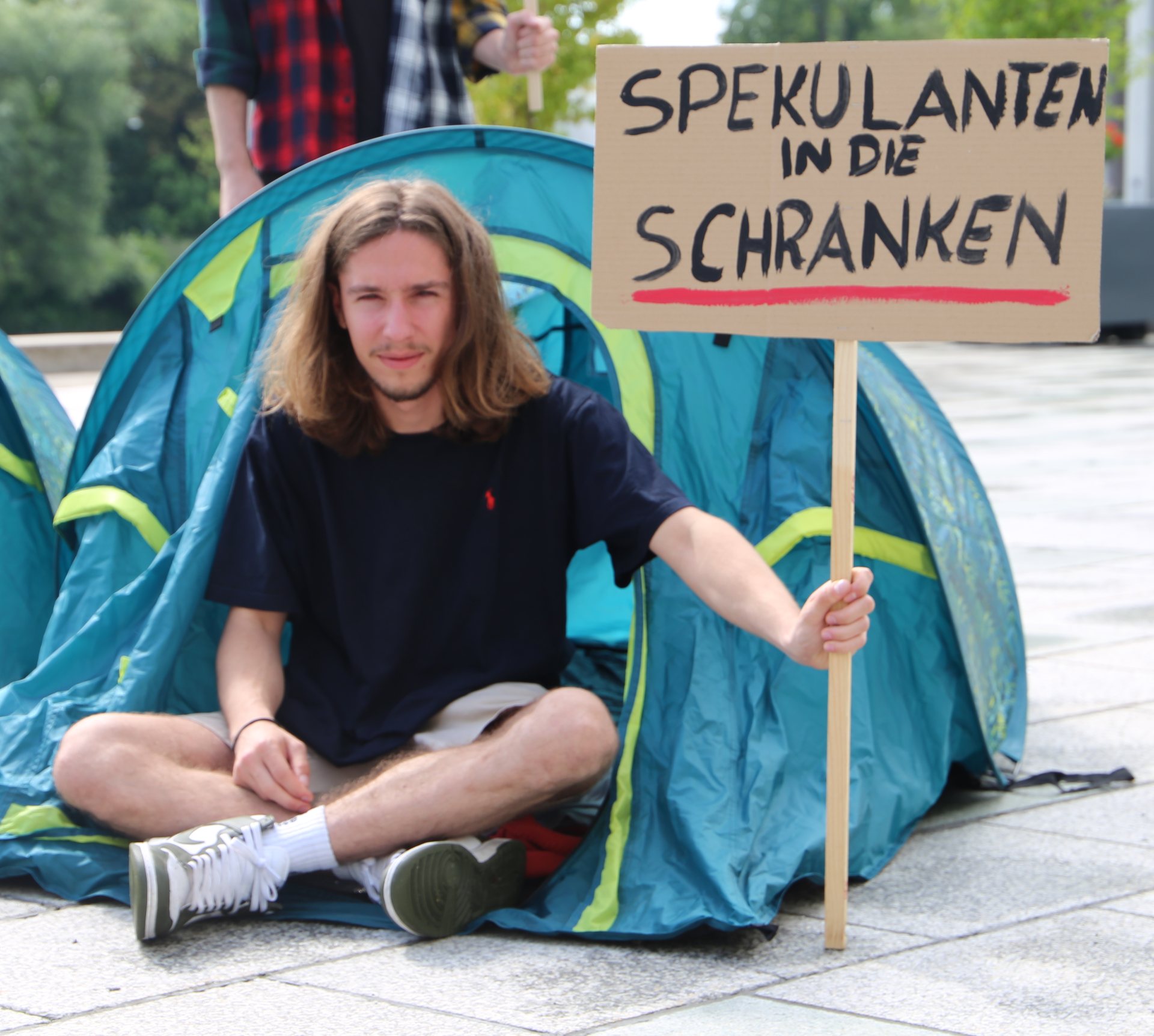 Ein Mann sitzt vor einem Zelt und hält ein Schild. Auf dem Schild steht oben: "Spekulanten in die Schranken".